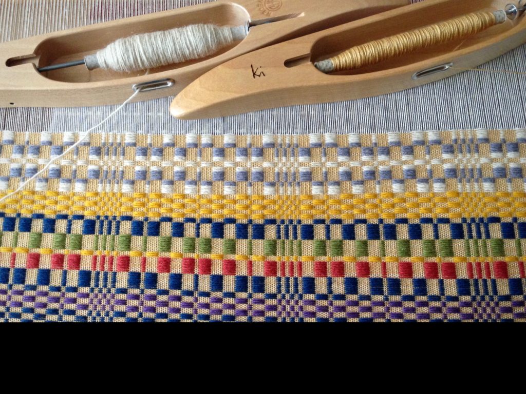 Swedish overshot, such as monksbelt, uses two shuttles--one for fine thread, and one for the thicker pattern weft. Warp is 16/2 cotton. Ground weave weft is 16/2 cotton. Pattern weft is 61 Fårö wool. Sett is 22 1/2 ends per inch. Weft density is 30 pattern picks per inch, with 2 tabby picks in between.