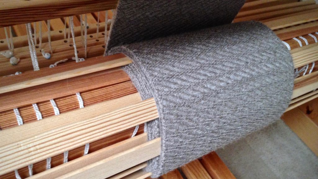 Woven fabric at the cloth beam. Warping slats between the scarves keeps the unwoven yarn (for fringe) from slipping off the cloth layers at the sides.